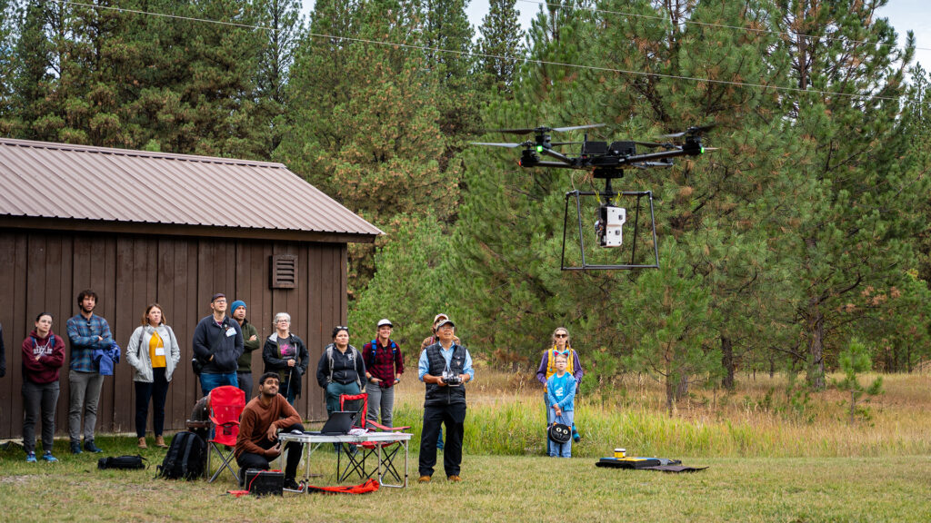 a group of people watch as a drone carry optical sensors is launched in a forest