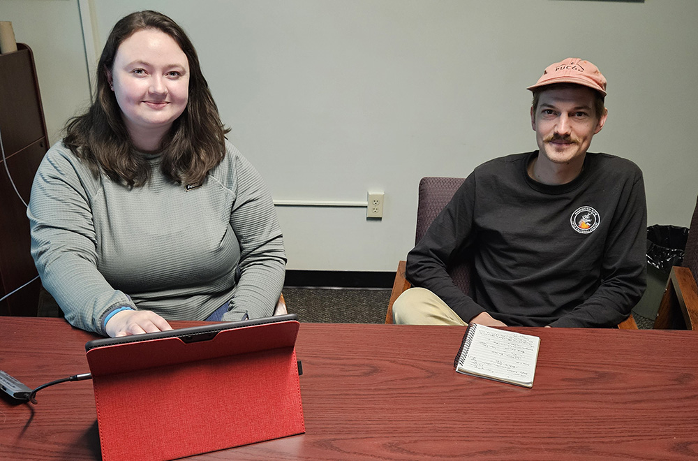 a woman works at a laptop and a man is seated to her left. Both are looking at the camera.