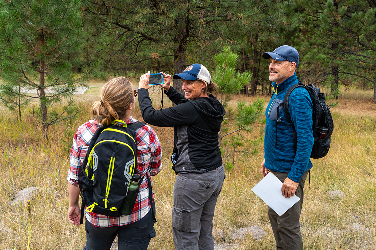three people in a forest with the woman in the center holding her phone up as to take a photo of the forest