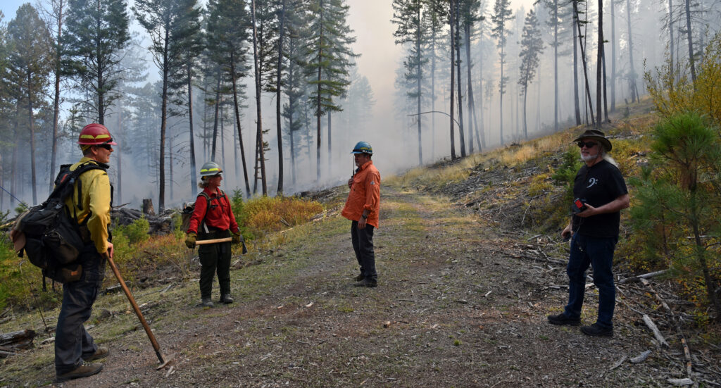 two fire crew members and other researchers talk in the road as a prescribed fire smoulders behind them