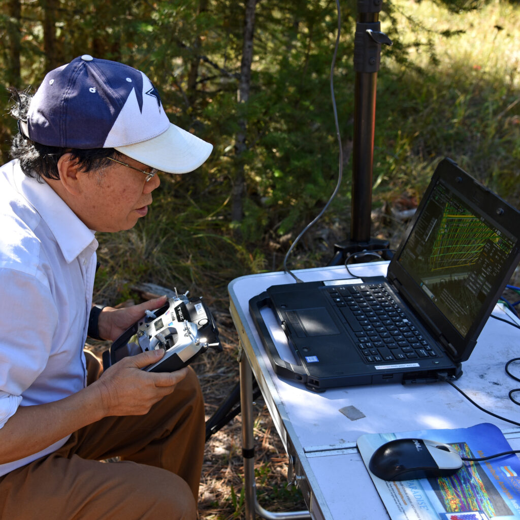 a man uses a controller to pilot a UAV (off-camera) while watching it's pattern on a laptop in a forest setting.
