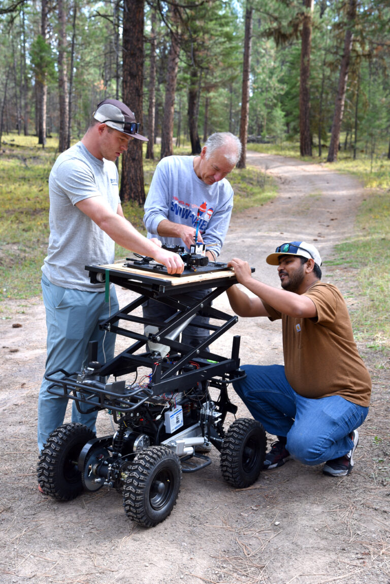 three people are adjusting sensors atop a ground rover in a forest